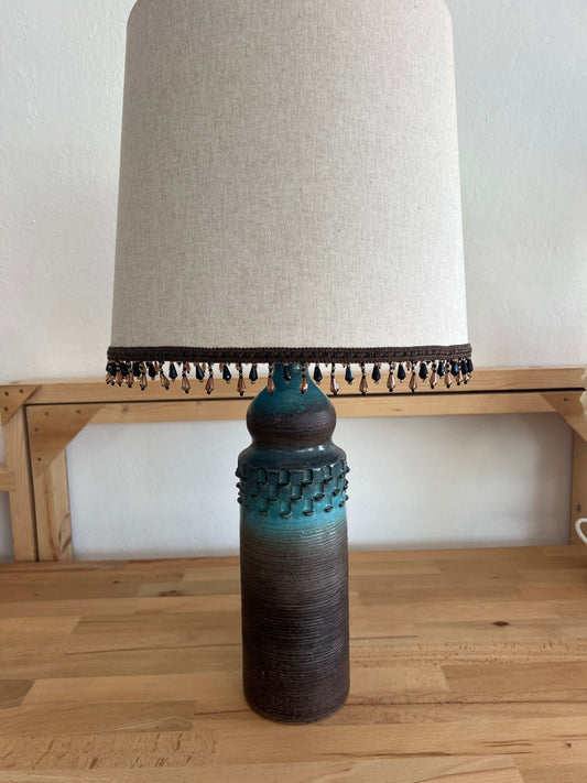 A handmade oatmeal beige lamp shade with a sky blue lining, featuring beaded detailed embellishment at its base, designed to fit a table lamp.