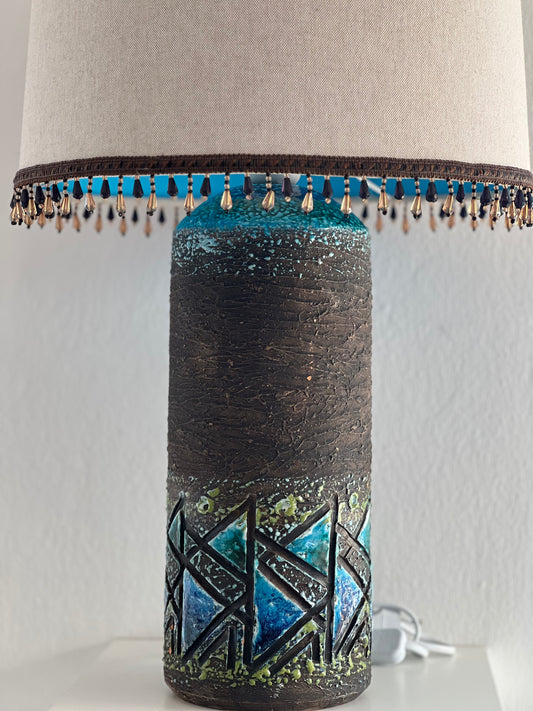 A ceramic table lamp with a geometric design, glazed in blue and green, displayed with a lampshade and a beaded trim on the base.